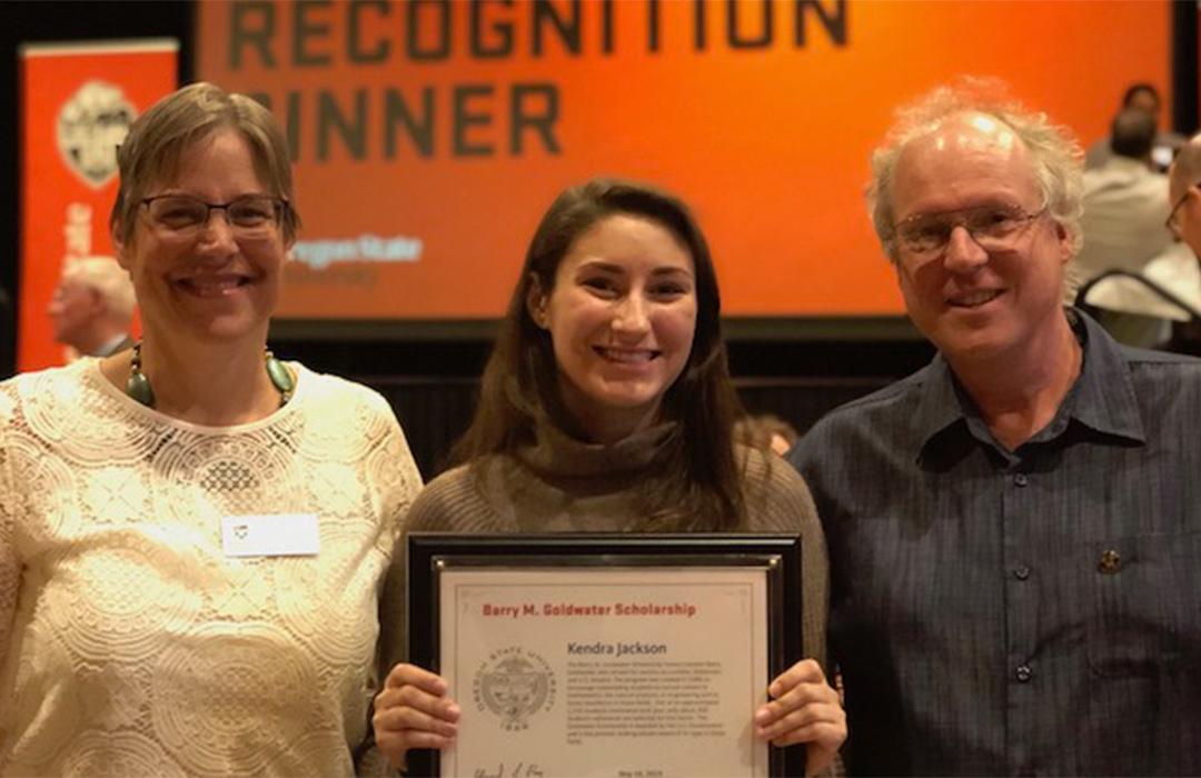 Kendra holding award with Kari Van Zee and Michael Freitag in auditorium