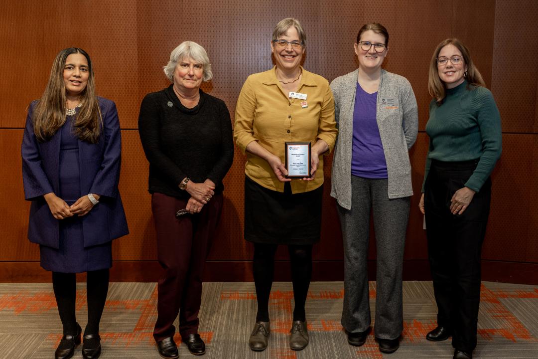 Five women pose for a picture. The woman in the center is wearing a gold shirt and holds a plaque.