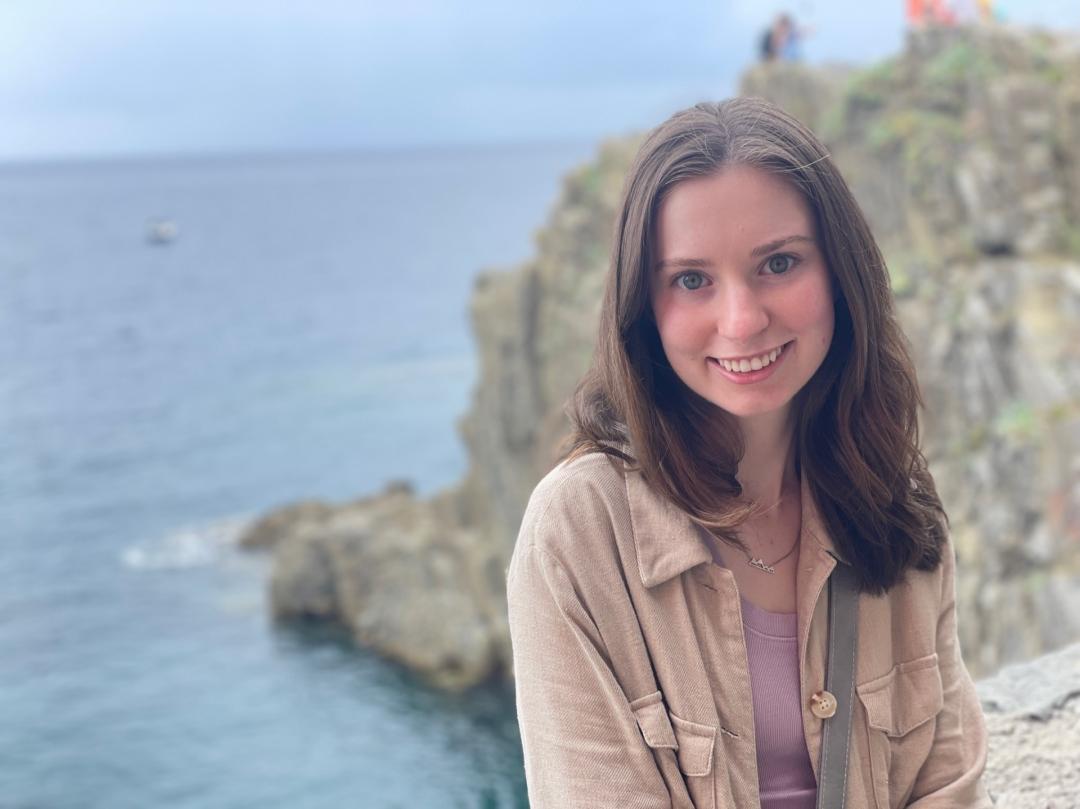 A woman stands with a wide smile in front of beige, rocky cliffs, which overlook a vast body of water.