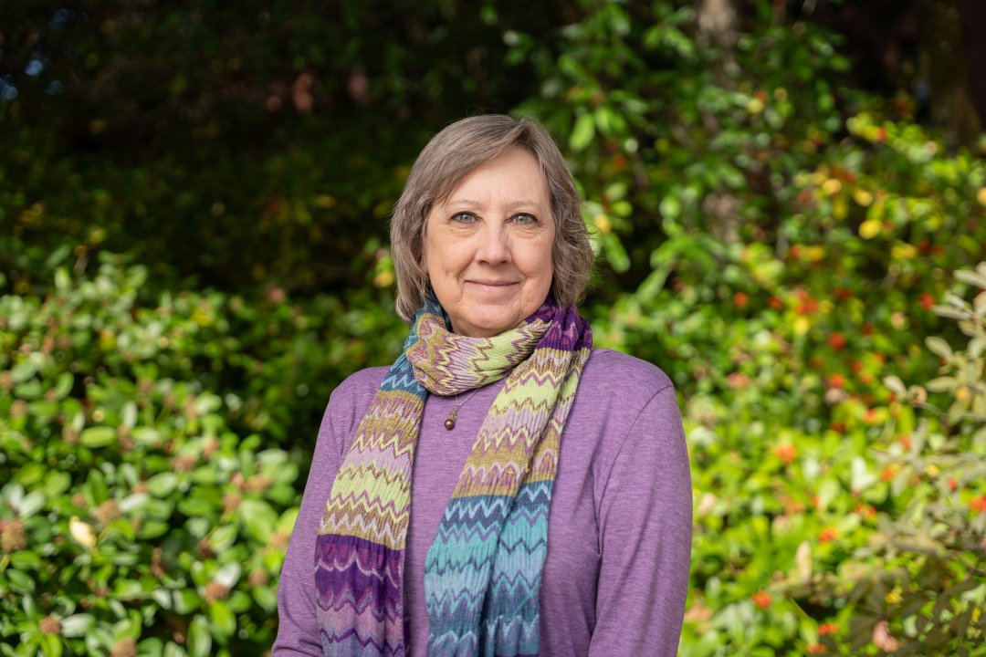 Bettye Maddux smiles slightly. She stands in front of lush greenery wearing a lavender sweater and colorful scarf. She has chin length gray hair.
