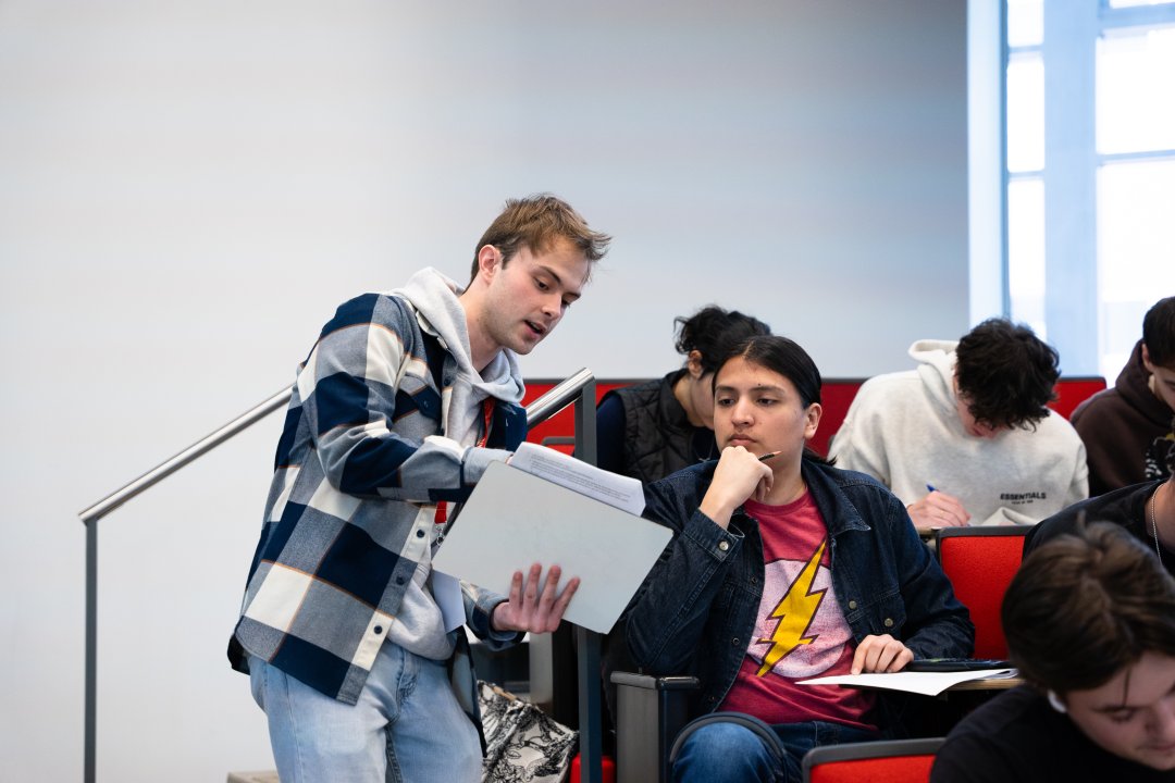 A man in plaid holds up a white board to a seated student, who listens intently as he explains a concept.