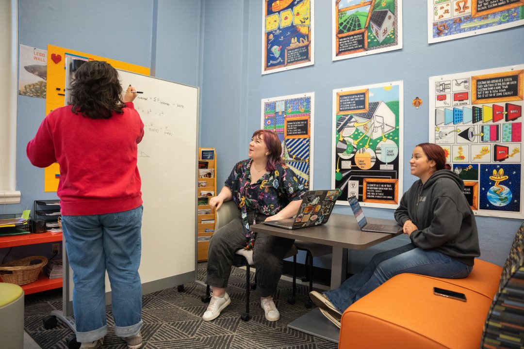 Three female students using a portable whiteboard to study chemistry together in a room with soft blue walls and science illustrations. 