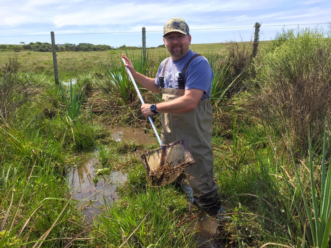 Jason Podrabsky smiling and holding fishnet standing in marshy field