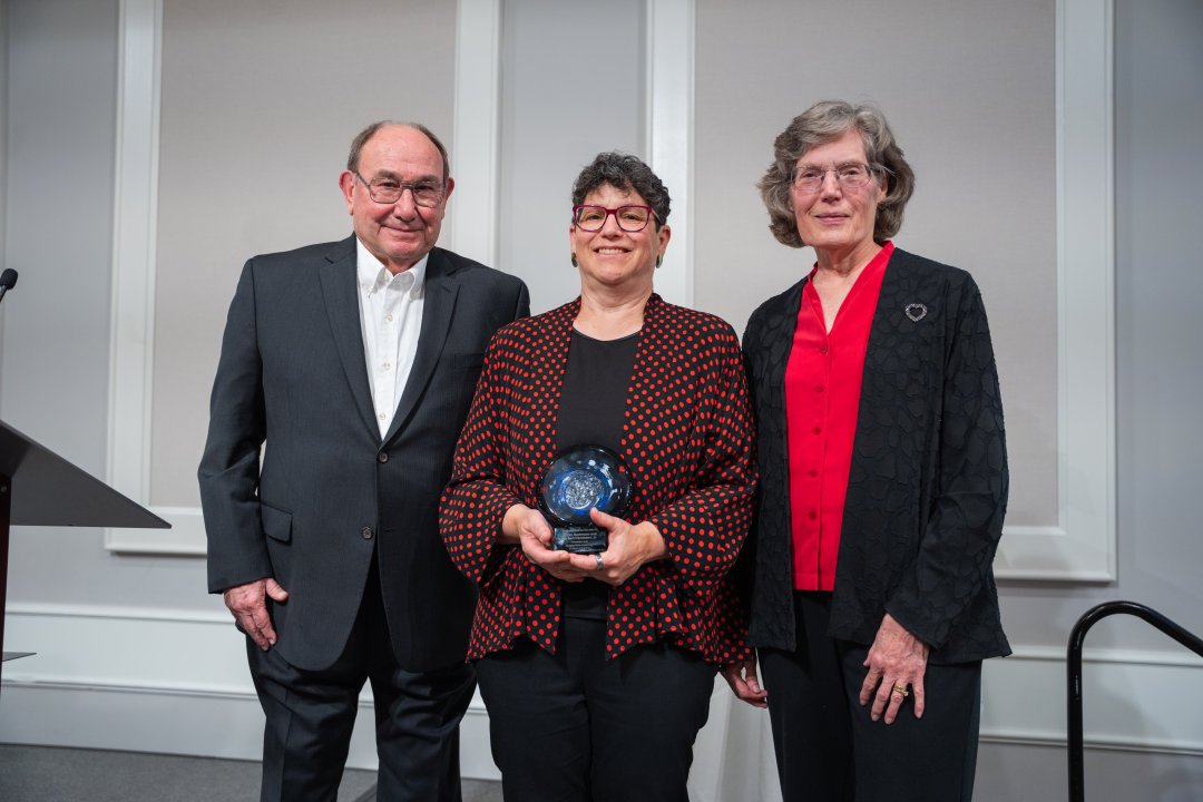 A man in a suit stands with two women in dress clothes. One of the women is holding a glass award.