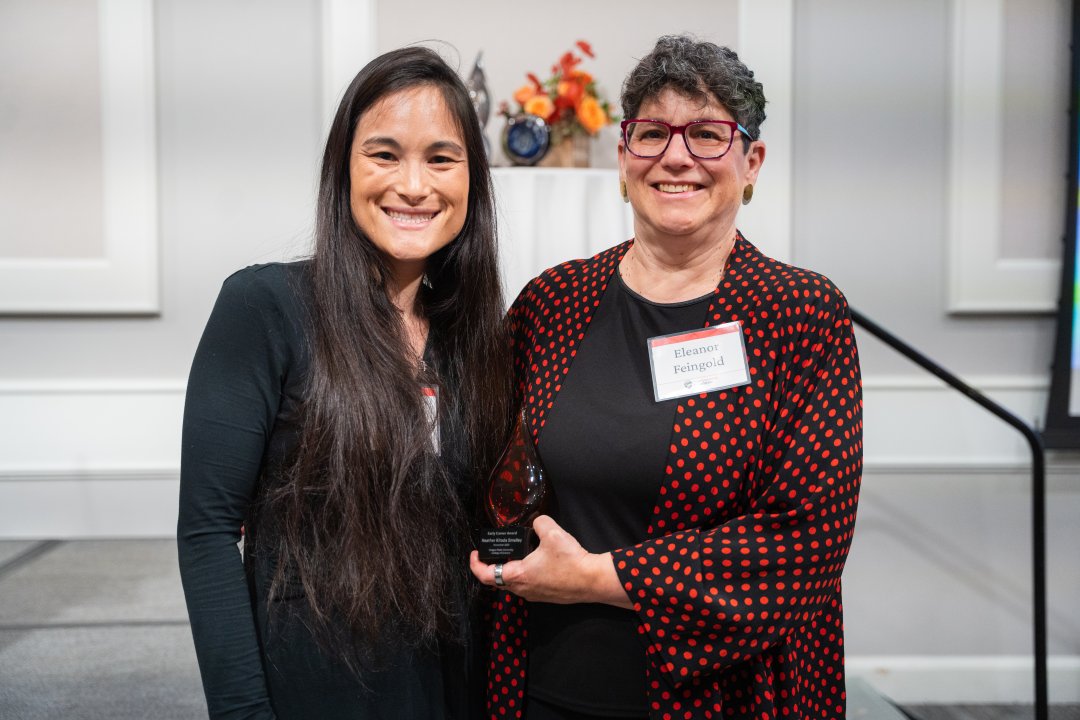 A woman in a black dress accepts an award from a woman in a black and red dress shirt.