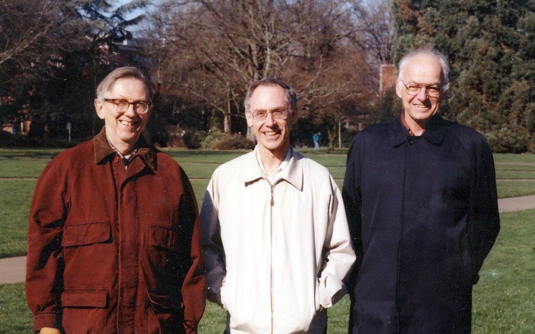 Three men smiling at the camera, wearing brown, white, and black jackets against an autumnal background on campus.