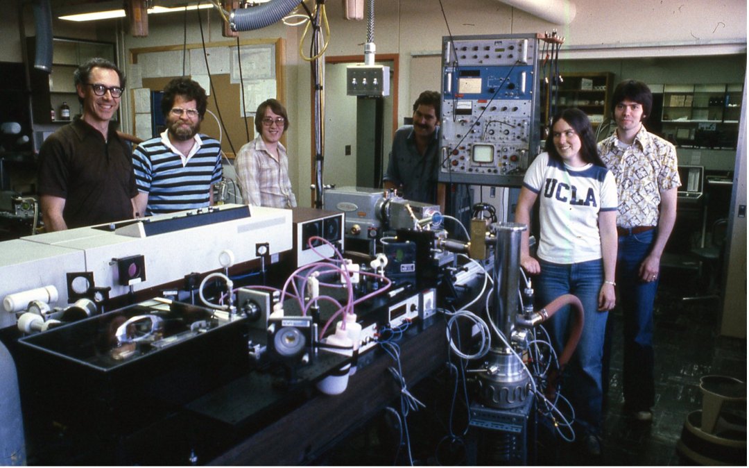 Several individuals stand around a large, wire-covered apparatus in a lab, dressed in 70s clothes.