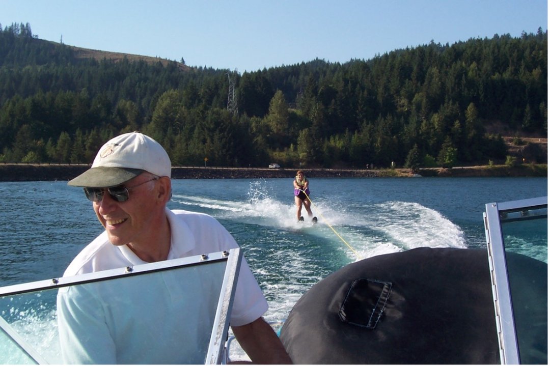 A man in sunglasses smiles as he drives a boat across a lake, with another person water skiing from the boat in the background.