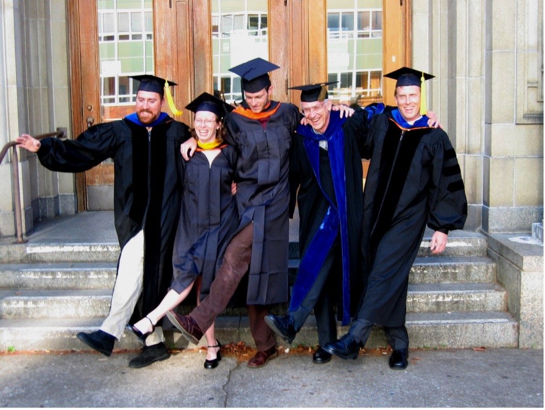 Five individuals don graduation regalia, smiling with their hands over each other's shoulders in front of a chemistry building.