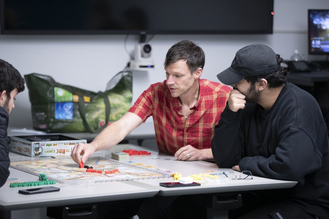 Two students and a professor sit a table and play the board game ticket to ride.