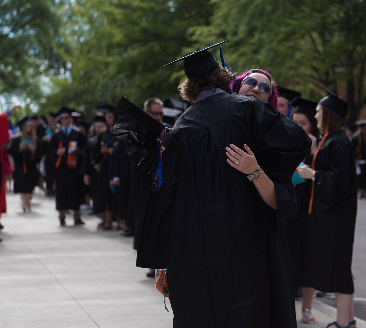 Two graduating students hugging each other in front of line of graduates in gowns
