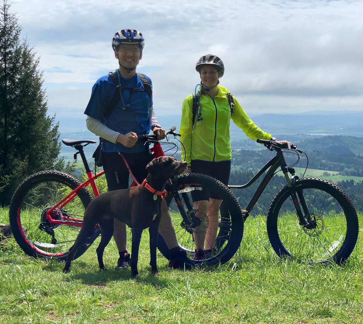 Courtney Rae Armour with husband and dog riding mountain bikes atop grassy hill