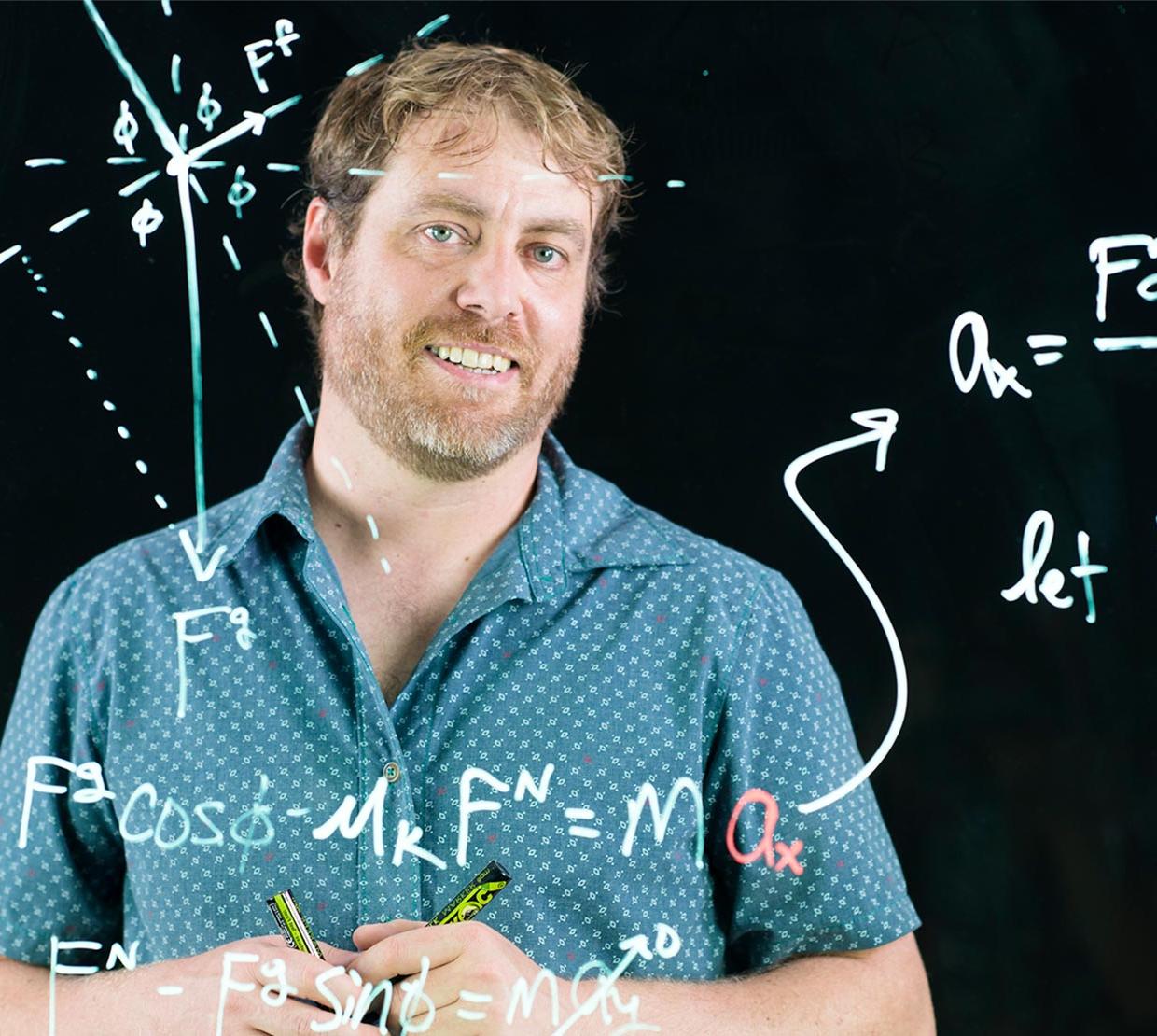 KC Walsh, writing out equations on window board with black backdrop