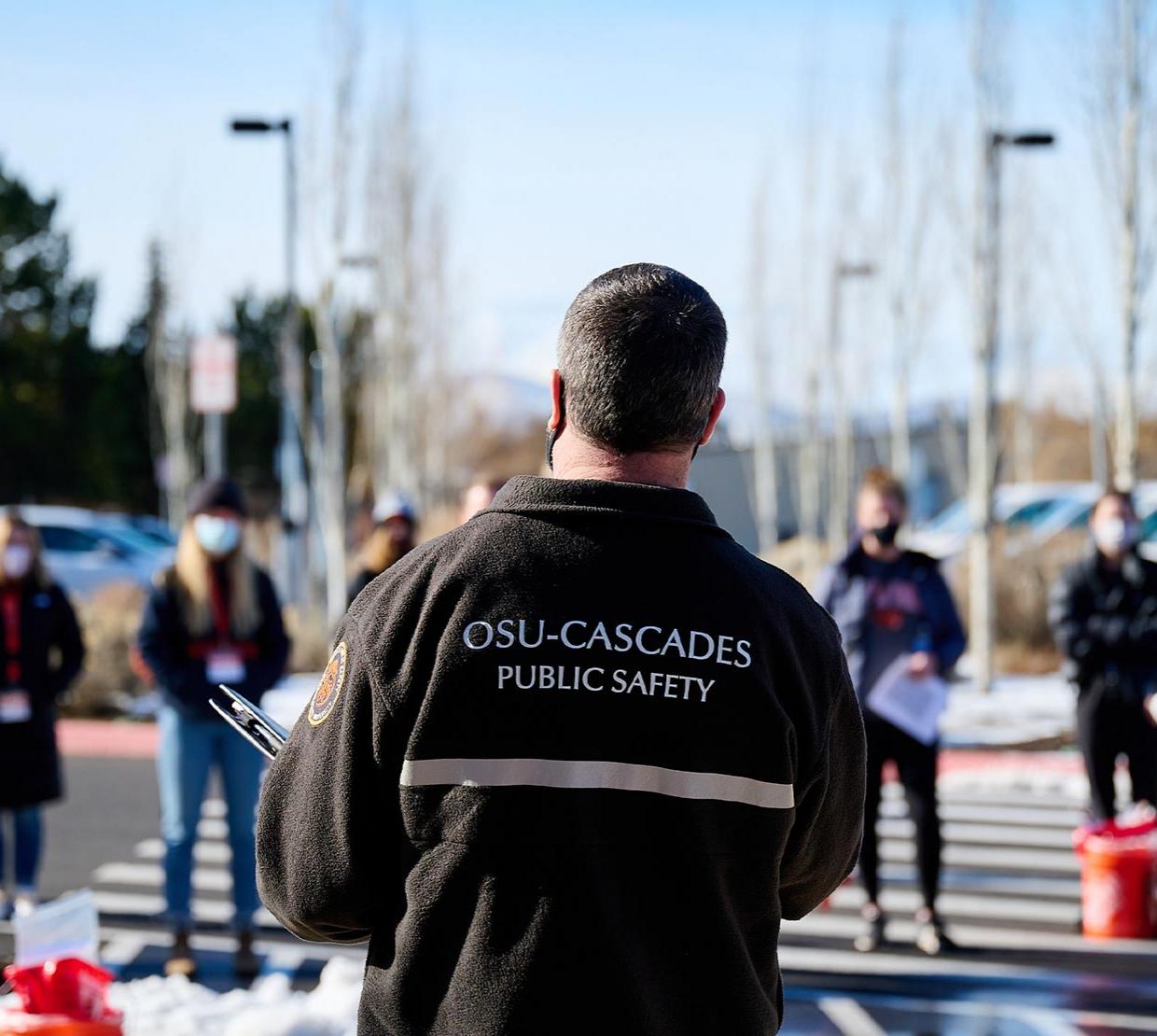 TRACE field staff member with his back to the camera, text on jacket "OSU-Cascades Public Safety" talking to and working with TRACE field staff members in snowy parking lot in Redmond, Oregon.