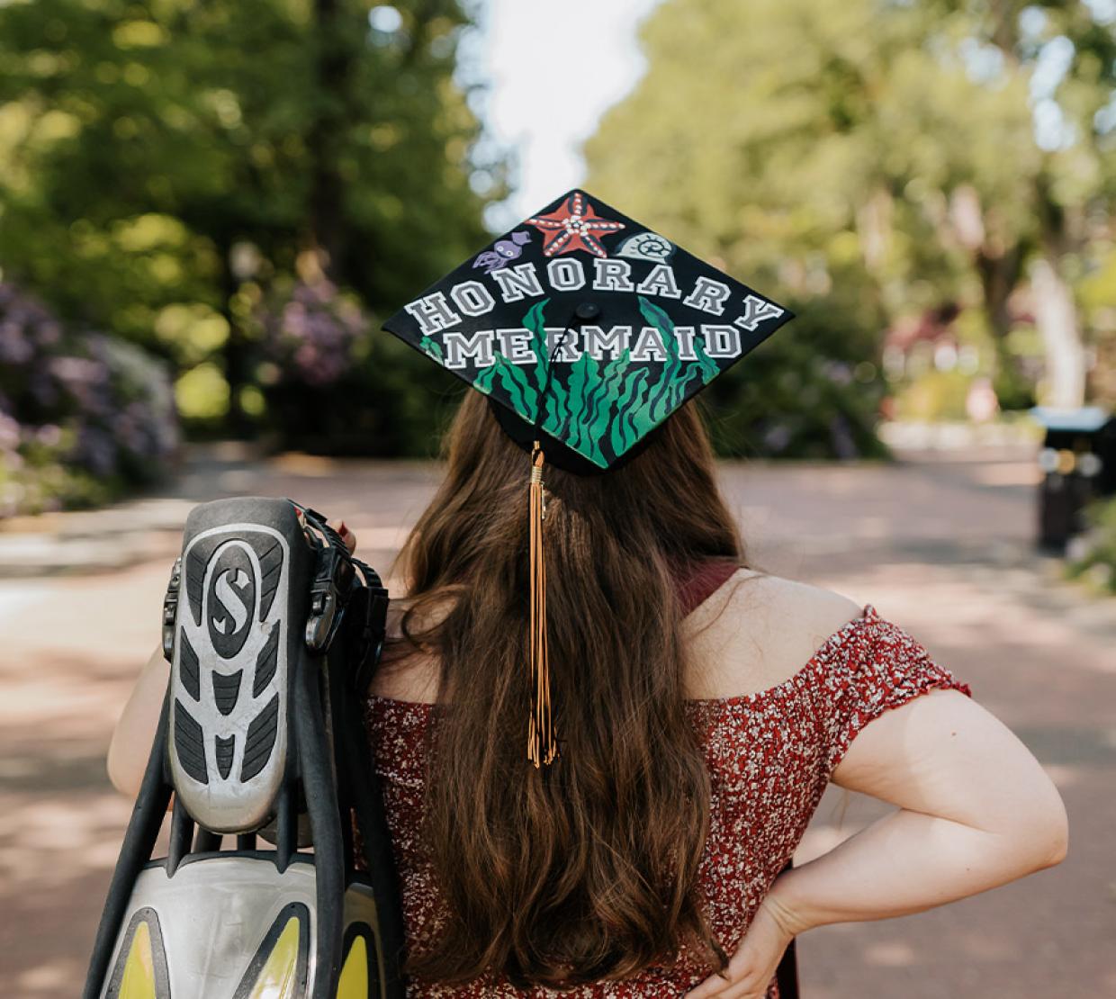 Simone Burton facing away from the camera, holding her scuba fins and wearing her decorated grad cap that says "Honorary Mermaid".