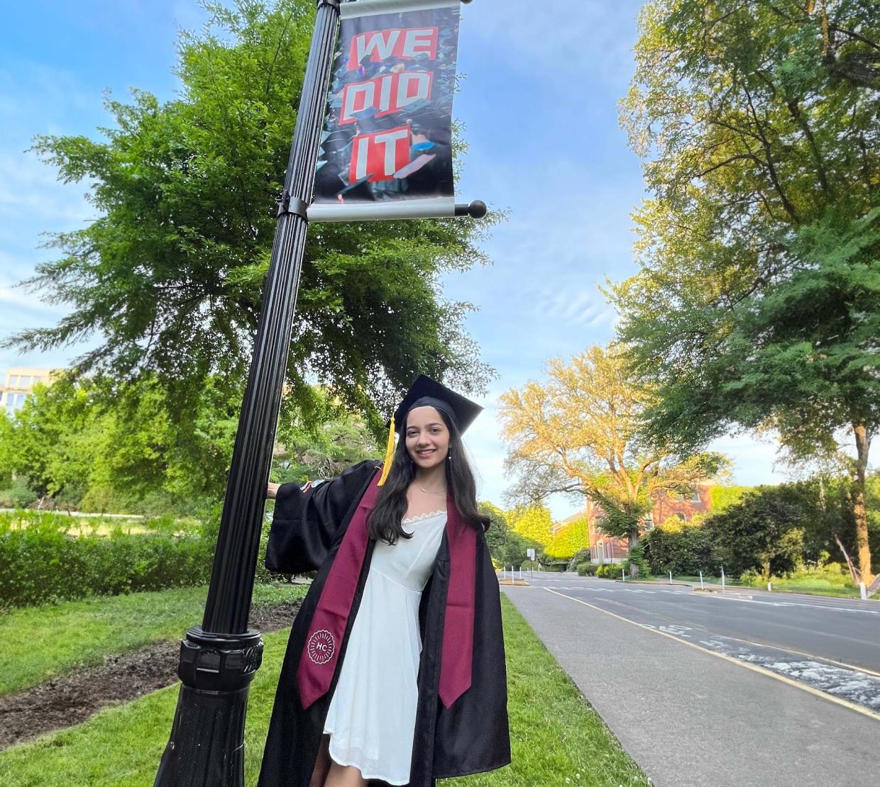 Alyssa Pratt stands in front of a pole with a sign that reads "We did it." She is wearing her graduation cap and gown.