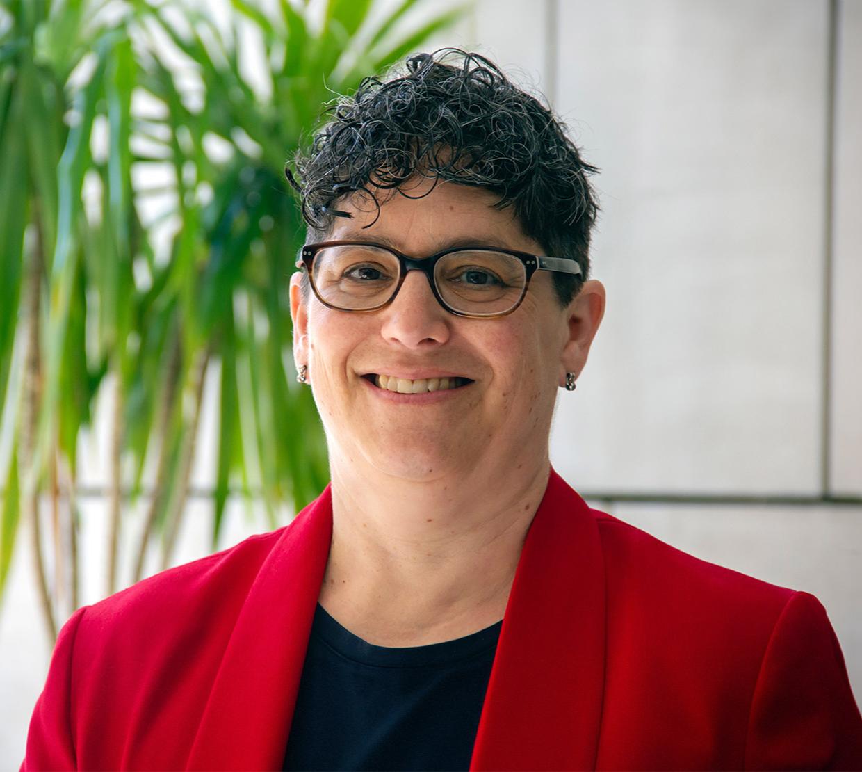A woman with short dark hair poses for a headshot wearing a black shirt and red suit jacket.