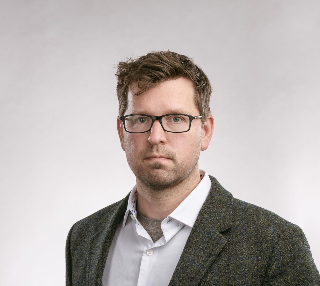 A headshot of a man in a grey suit with white shirt.