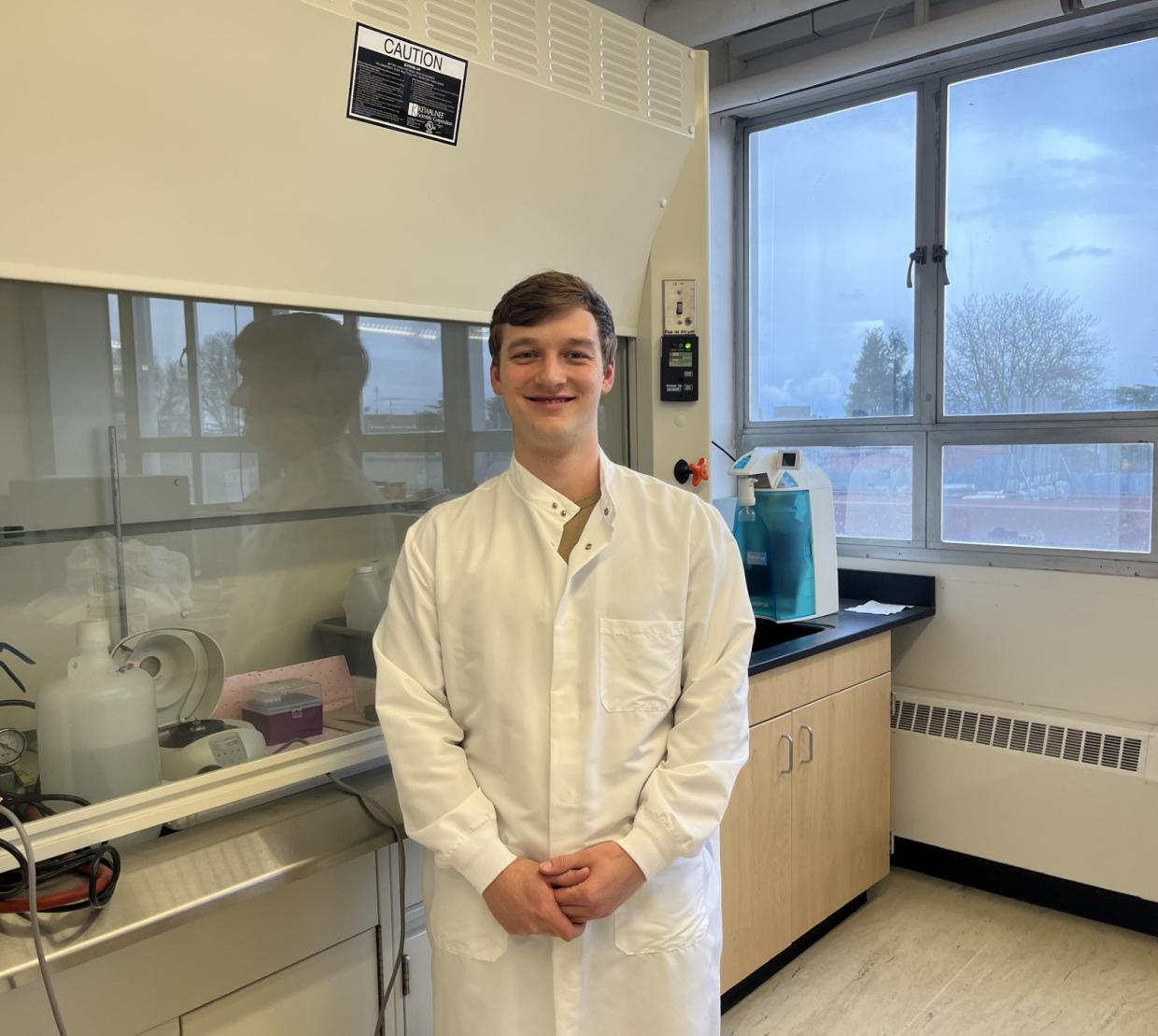 Kelly Shannon dons a white lab coat in an OSU laboratory, equipment lining the wall on his right and a window framing trees outside on his left.