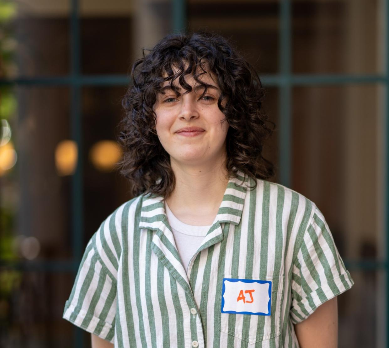 AJ Damiana wears a striped button-up with a smile, her dark, curly hair falling to her shoulders.