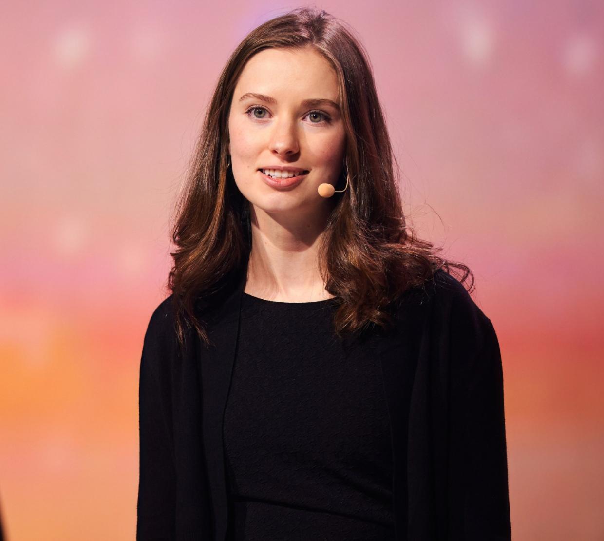 A woman with shoulder-length hair wears an earset microphone and presents to an unseen audience