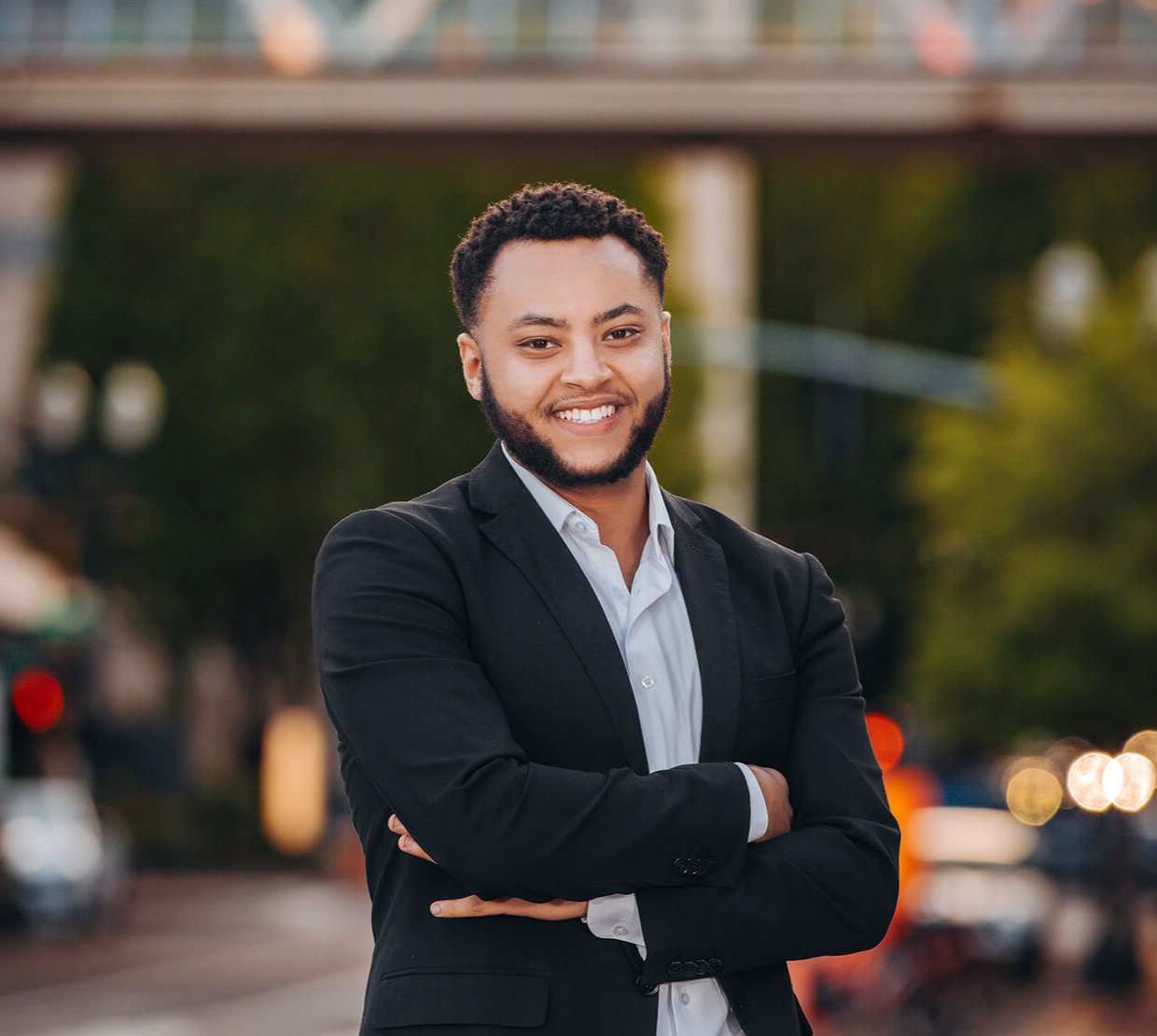 A man in a black suit stands in front of a blurry city background. 