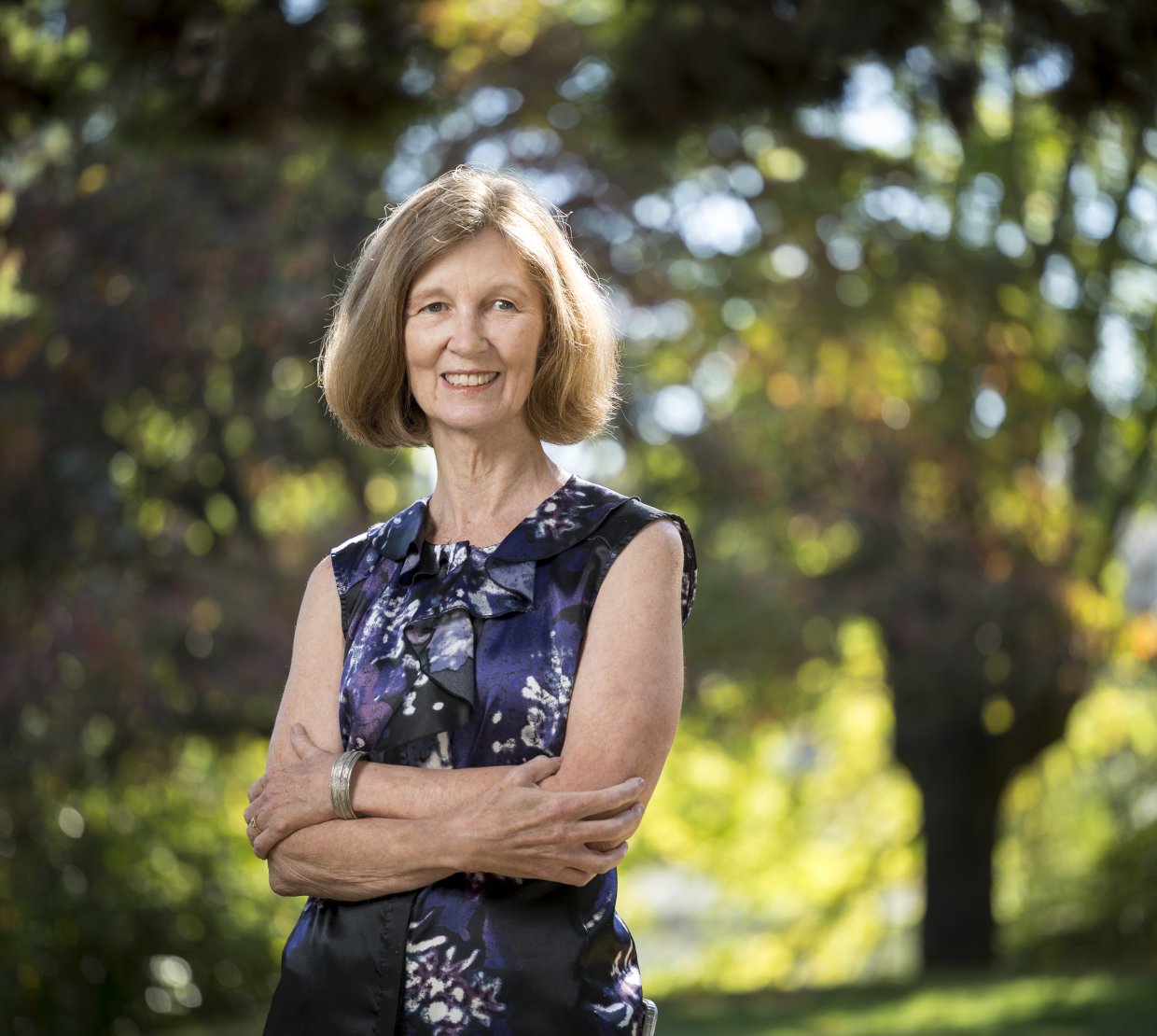 A woman with short hair stands with arms crossed in front of a green nature background.