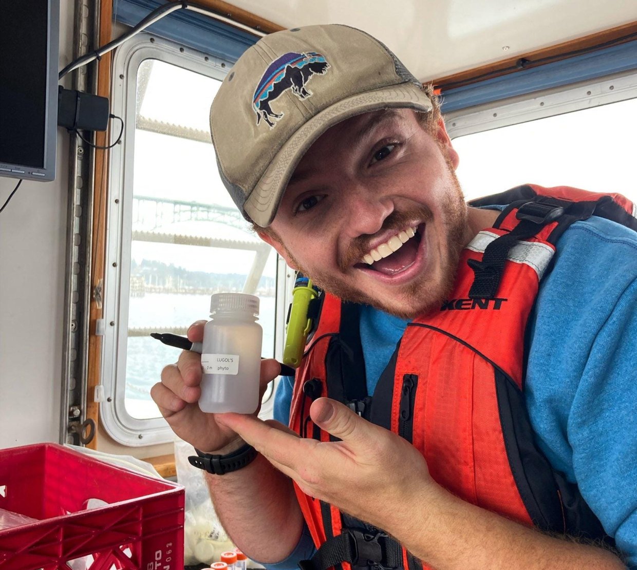 A man with a slight beard wearing a tan hat with a bison on it, and wearing an orange lifevest, holds a sample bottle.