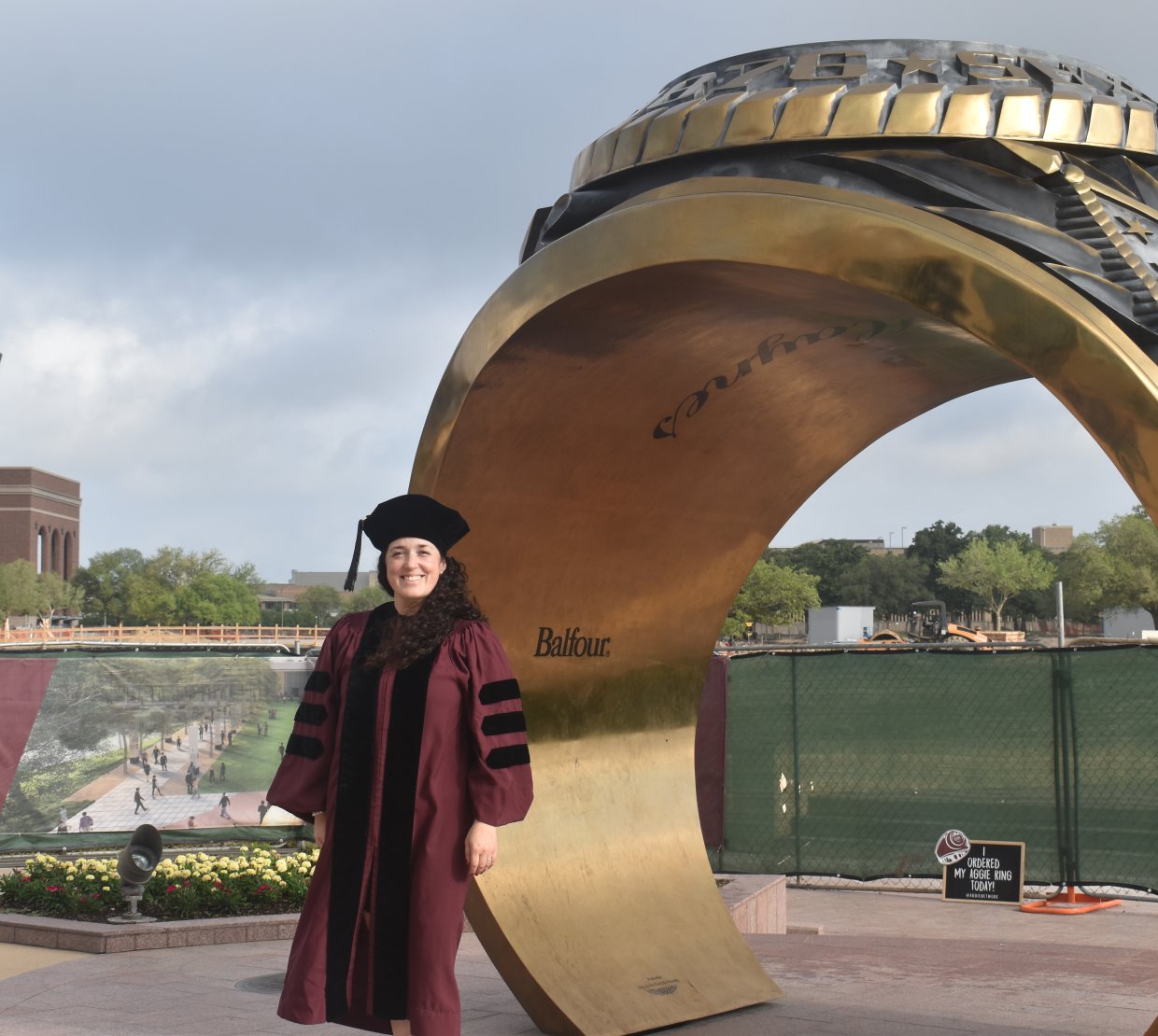 A woman wearing a deep red graduation gown stands on a college campus in front of a large golden ring statue.