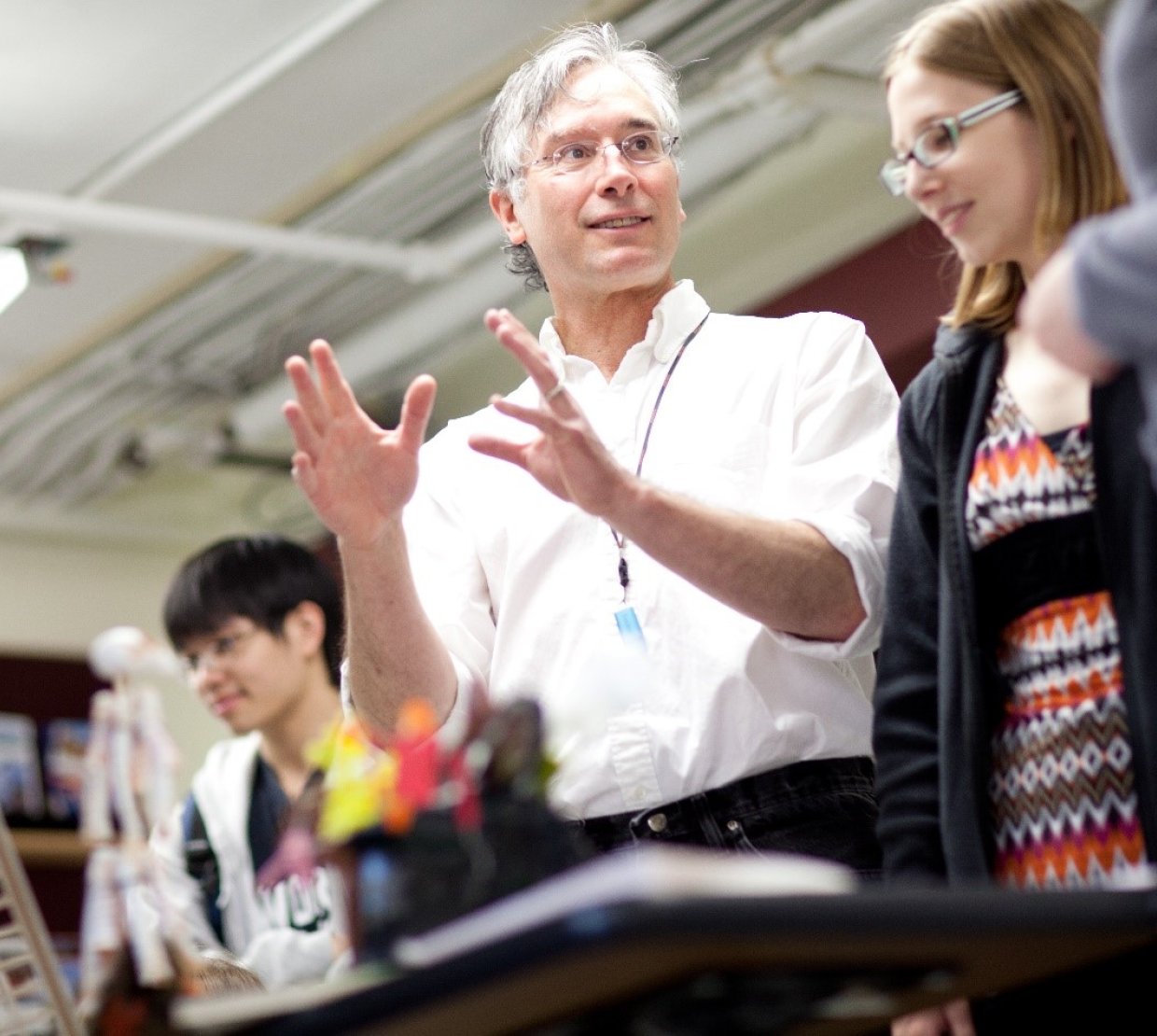 A man in a white button-up gestures to molecular art on a table, explaining it to onlooking students.