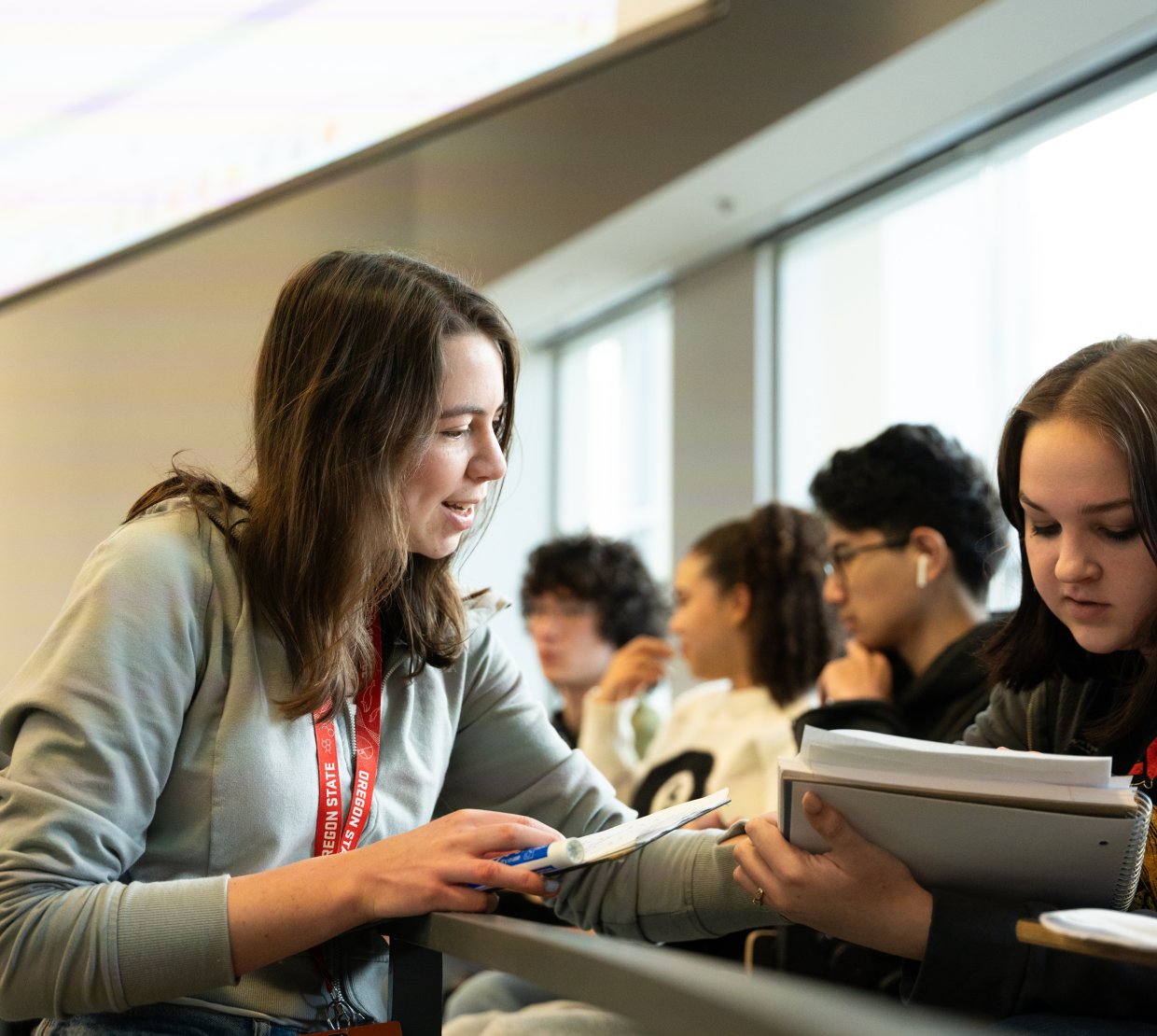 A woman leans over seat railing to aid a student, pointing to the student's notebook mid-conversation.