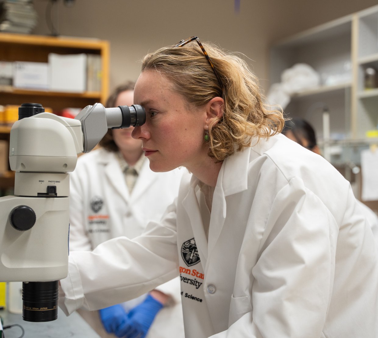 Two women in lab-coats look into a microscope.