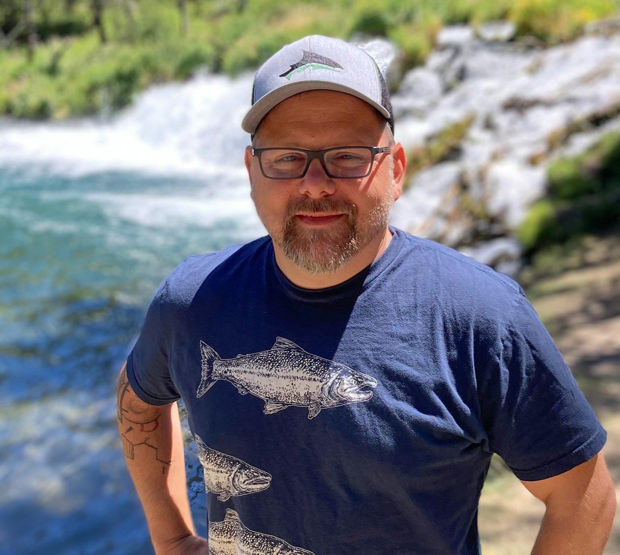 Jason Podrabsky in cap and fish shirt smiling in front of river