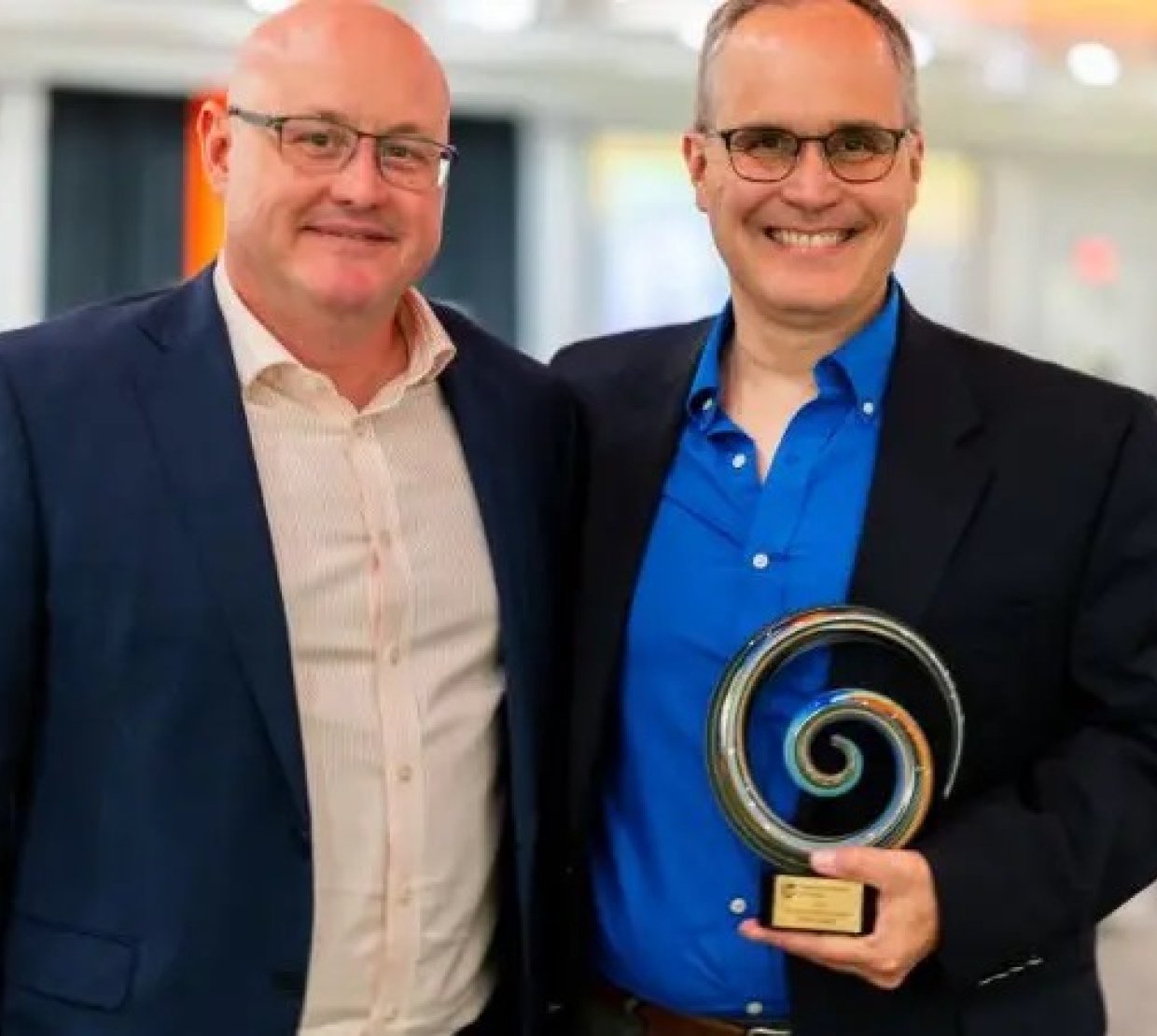 Two men stand wearing suits. One of them is holding an award.