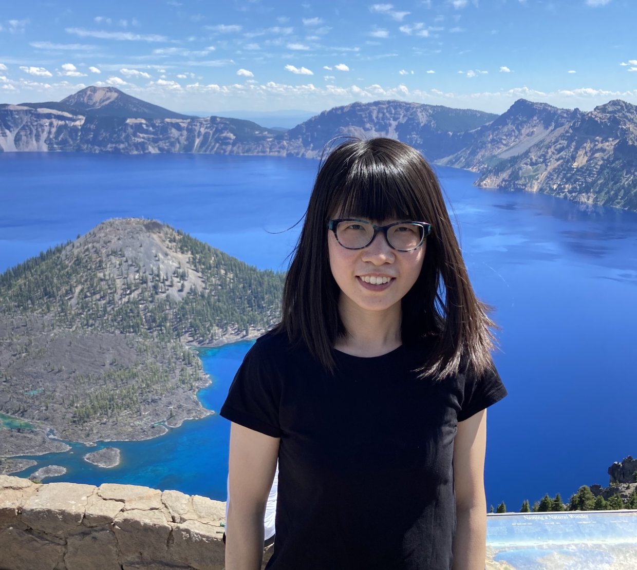 A woman in a black shirt poses for a photo in front of a stunning blue mountain lake.
