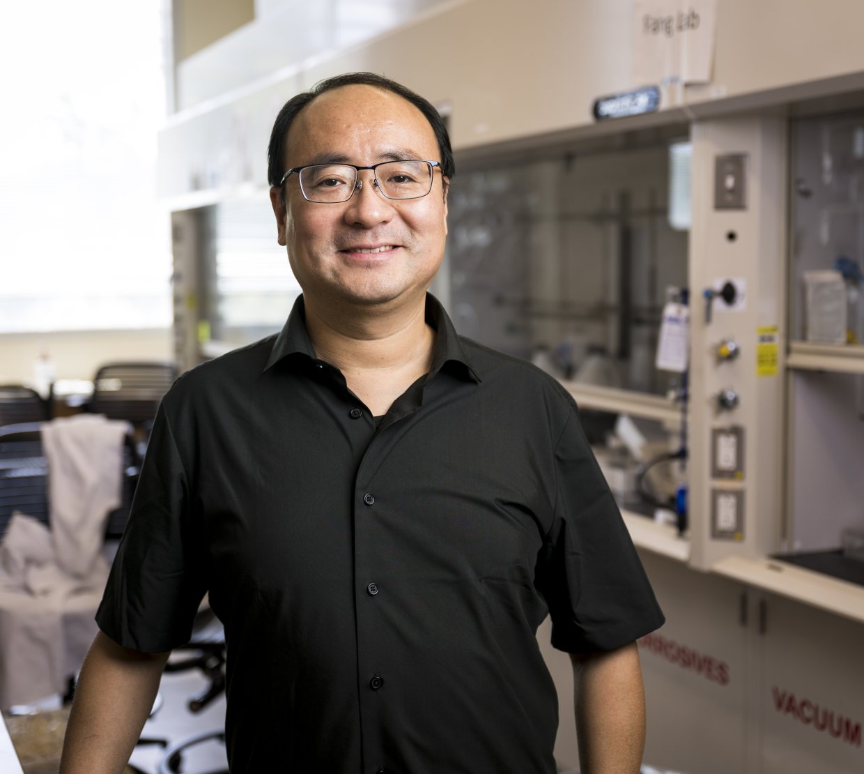 A man in a short sleeve black shirt smiles while standing in a laboratory. 
