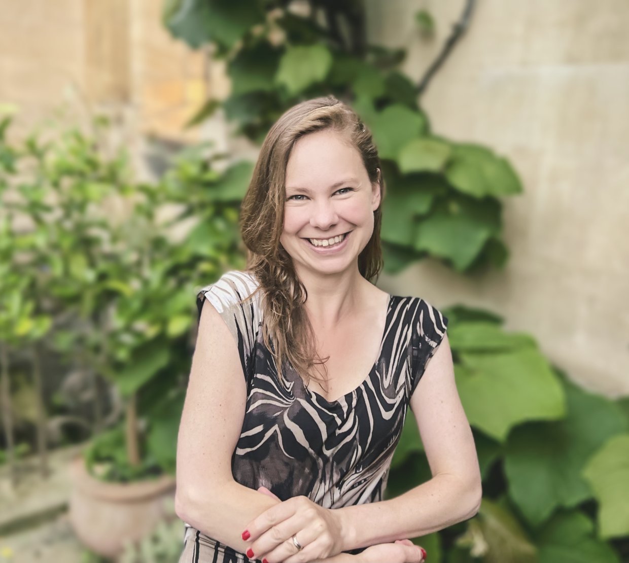 Headshot of Dr. Laura P. Schaposknik: A woman smiling with her hands folded in front of her, sitting in front of a background featuring beige stone walls and green plants.