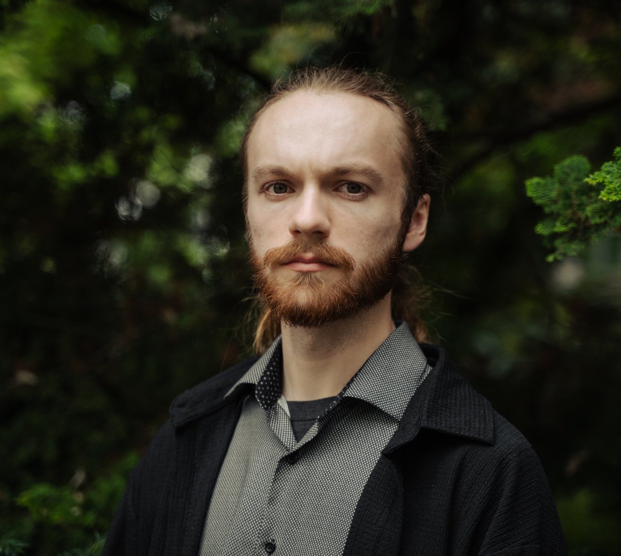 A man in a grey shirt and black coat poses for a photo in front of trees.