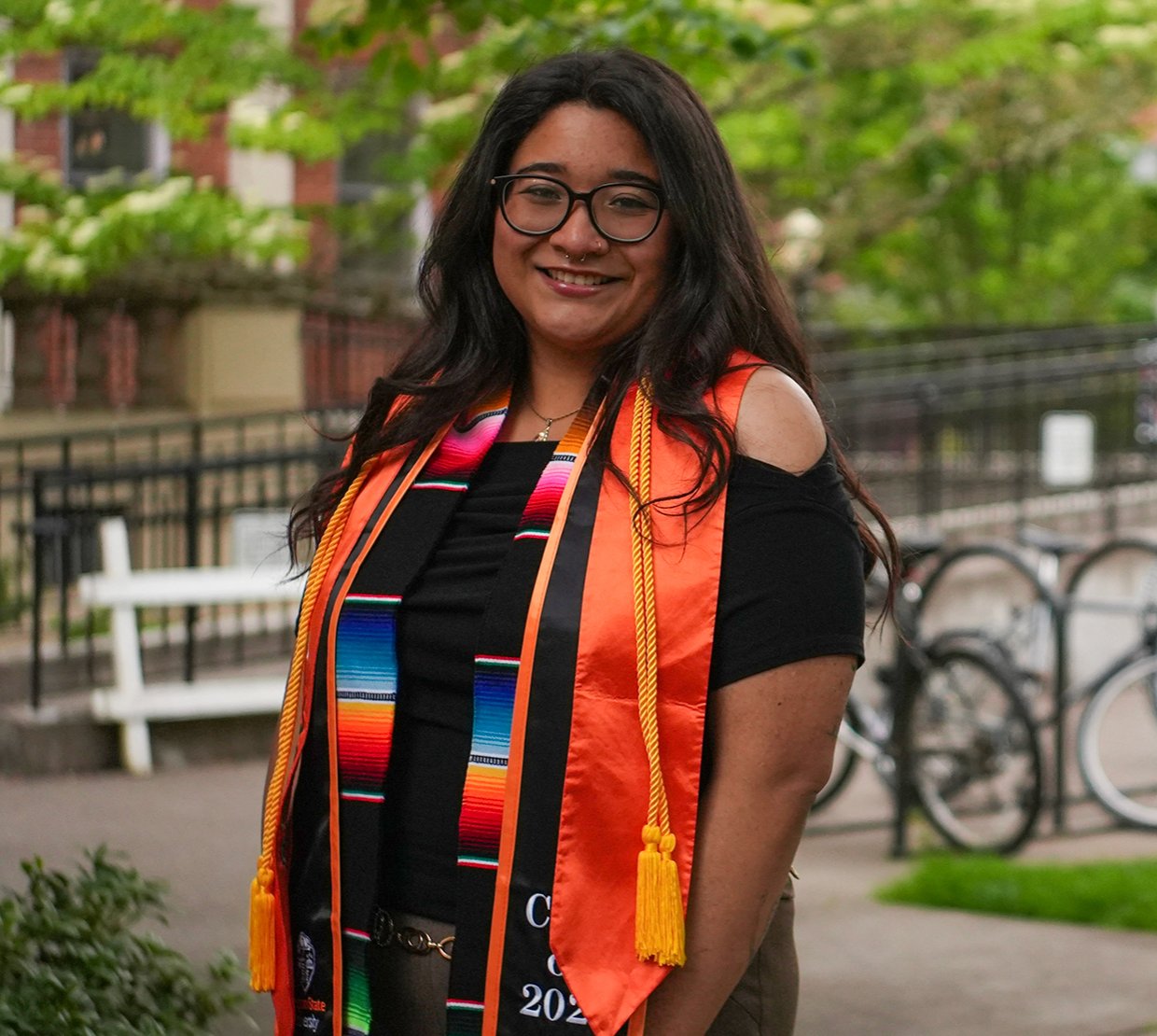 A woman in glasses and graduation regalia smiles brightly in front of a sunny campus backdrop.