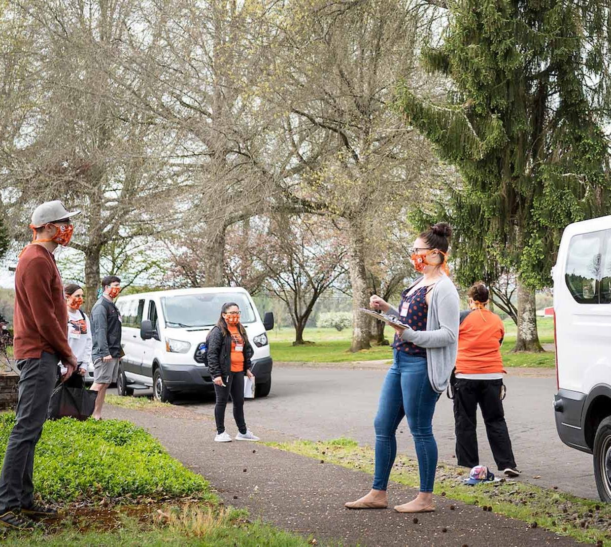 TRACE staff wearing face masks and face shields brief community volunteers before COVID-19 testing outside near two white vans.