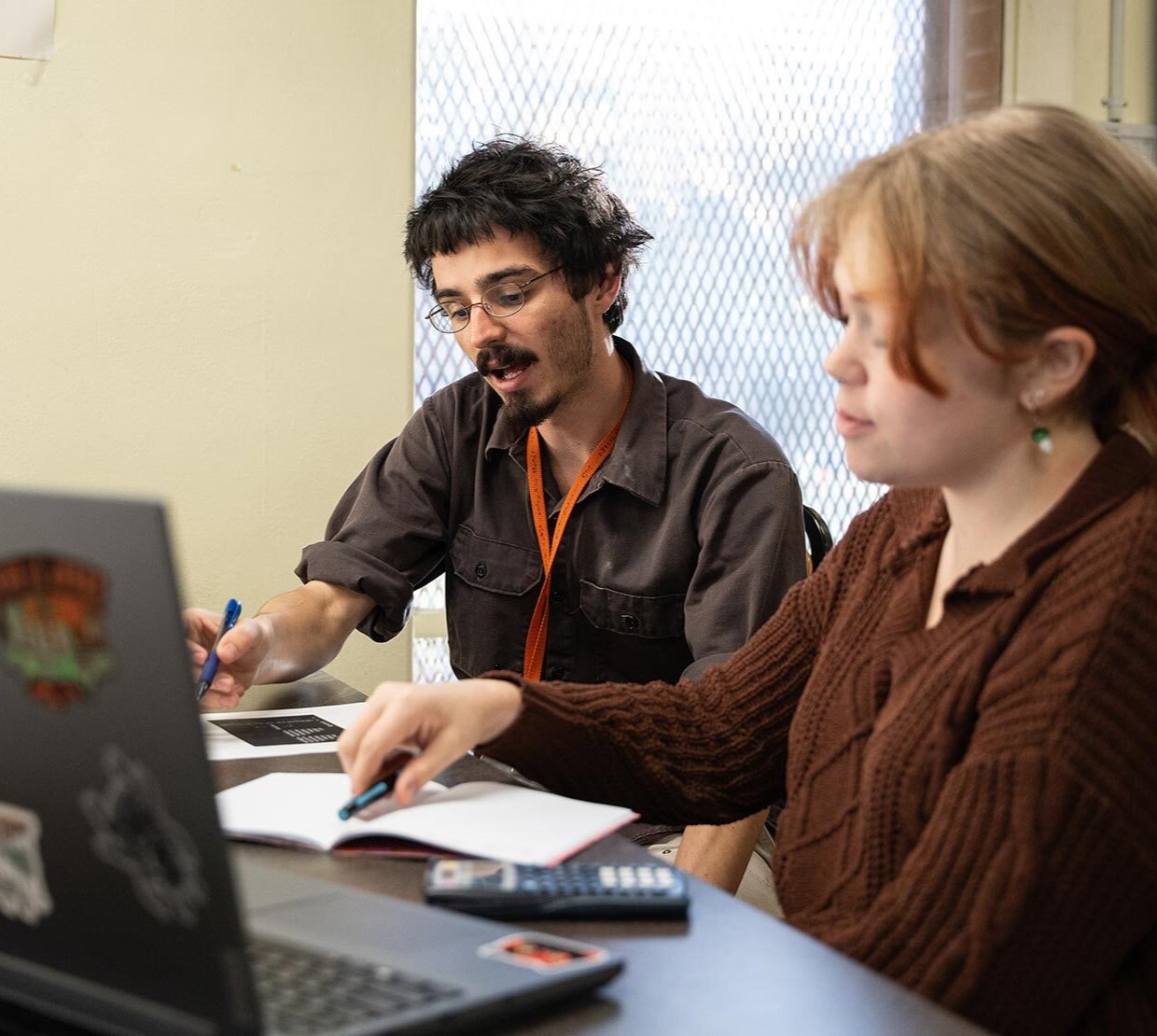 A teaching assistant sits beside a student at a table, pointing to coursework with a pen as they work together. An open laptop, notebook and calculator are on the table, and a MTH 112 sign hangs on the wall behind them.