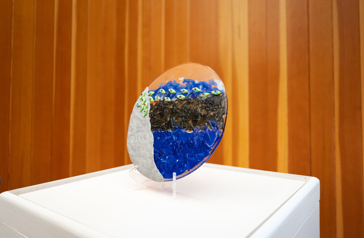 A glass art piece in Vaishnavi's show stands on top of a white museum pedestal. It is in shades of blue and black.