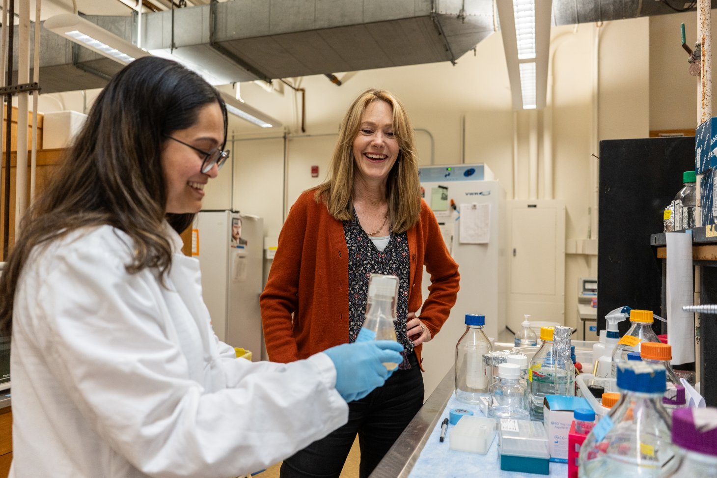In the lab at Oregon State, Vaishnavi Padaki stands in the foreground wearing a lab coat. She has long dark hair and wears glasses. She is holding a flask with liquid in it. In the background, you see her mentor, Dr. Kimberly Halsey wearing a floral top and red cardigan sweater. She stands with one hand on her hip, looking at the flask in Vaishnavi's hands. She smiles widely as though she's laughing. The bench in front of them is littered with various lab equipment.