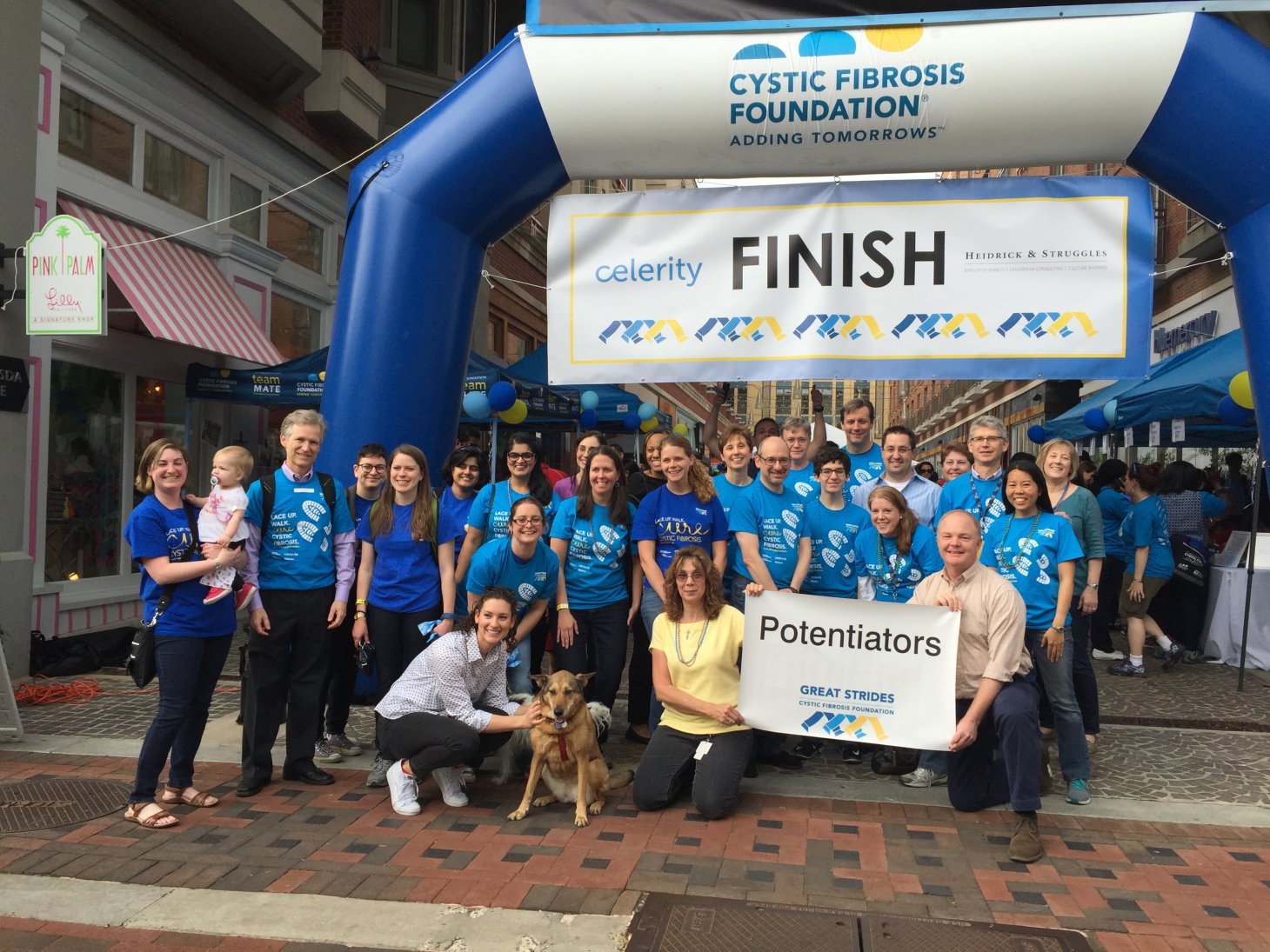 A group of people, including Skach, smile for the camera in front of the finish line. They are wearing blue shirts with white footprints as graphics. A couple members of the group hold a giant poster sign that says, "Potentiators."