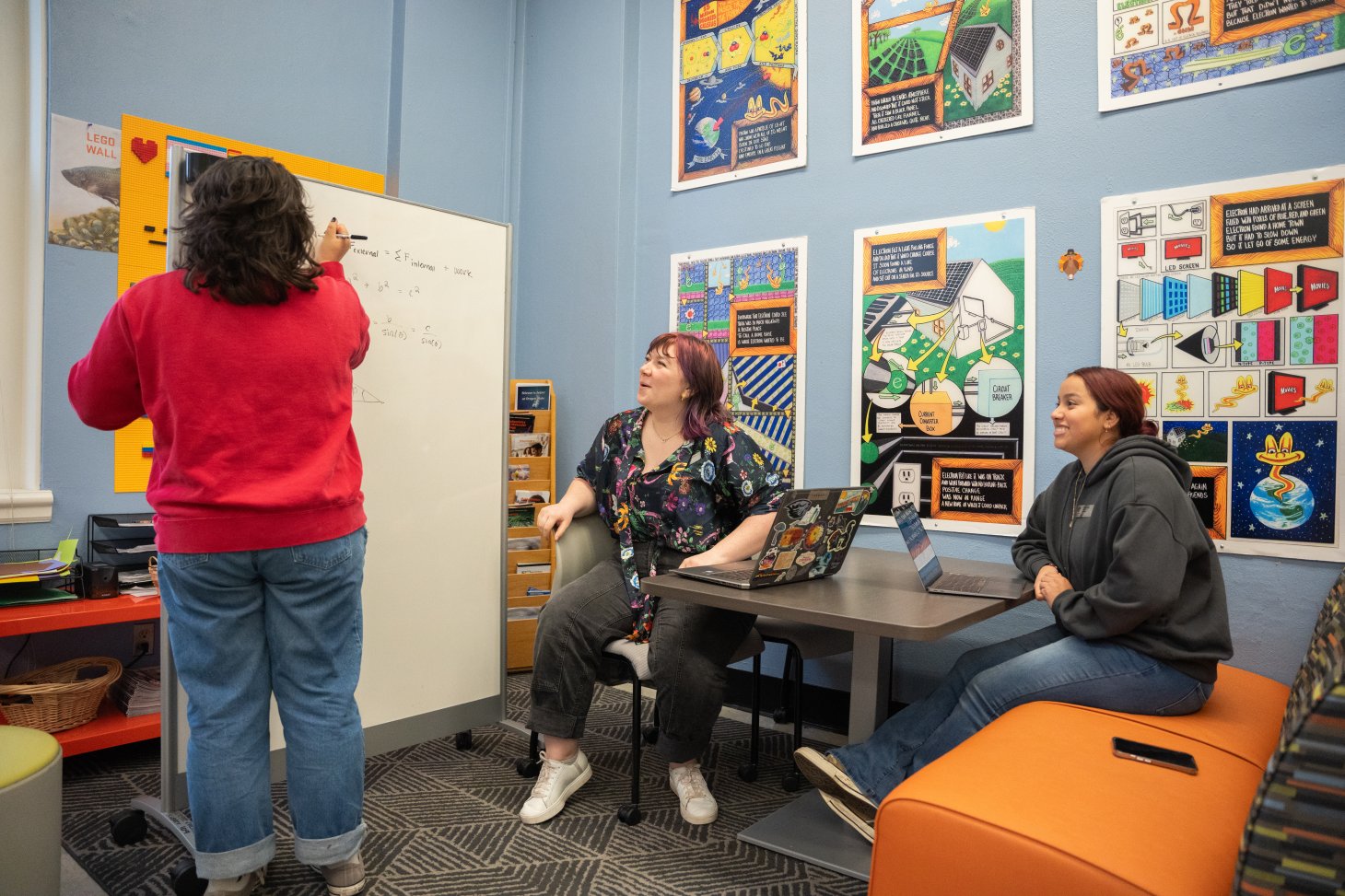 Three students collaborate in the Science Success Center. This is a colorful study space with large science posters on the wall. One student writes on a whiteboard. The other two sit at a table, viewing the board.