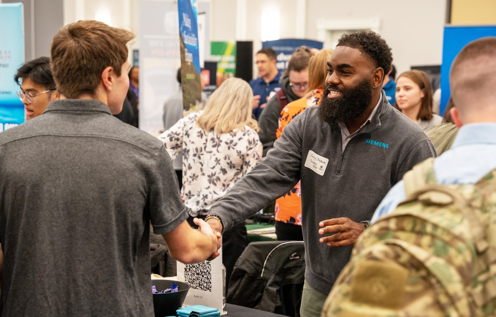 An industry professional smiles and shakes hands with a student across a table. Their shirt reads "SIEMENS."