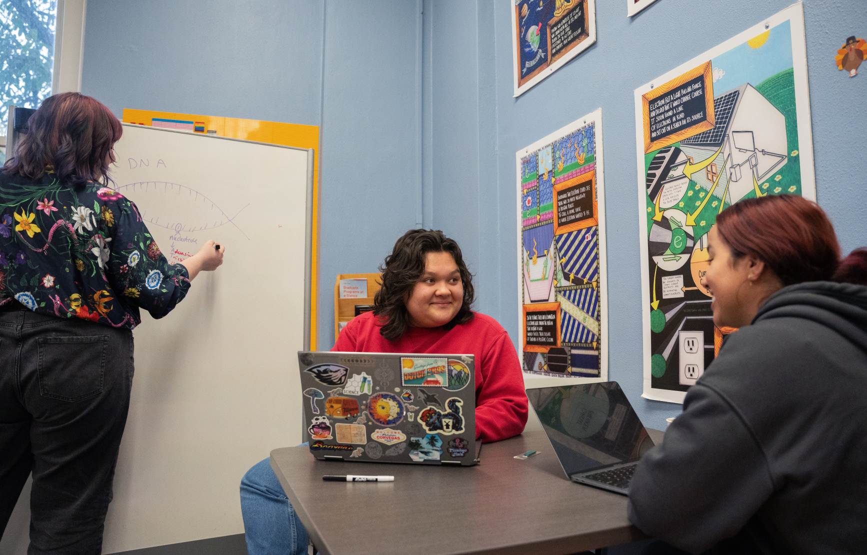 Three students work in the Science Success Center. Two are seated at a table, both with open laptops. The one in the center wears a red shirt and their laptop back is covered in stickers, including an OSU beaver. They smile at the student across from them. The third stands to the left of the frame at white board, drawing a DNA model.