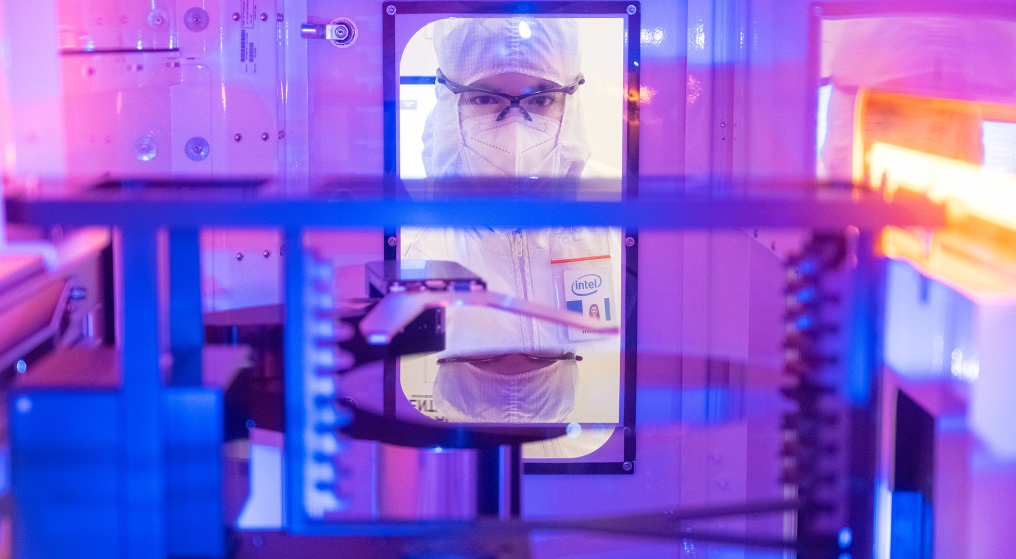 A woman in full PPE wearing an intel name badge peers through a small window in a piece of machinery.