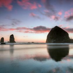 Three rocks jut up from Cannon Beach on the Oregon Coast during a sunset with pink clouds. 