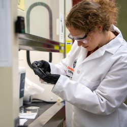 A student with her hair pulled back wearing a lab coat and protective goggles looks intently at the glassware in her hands.